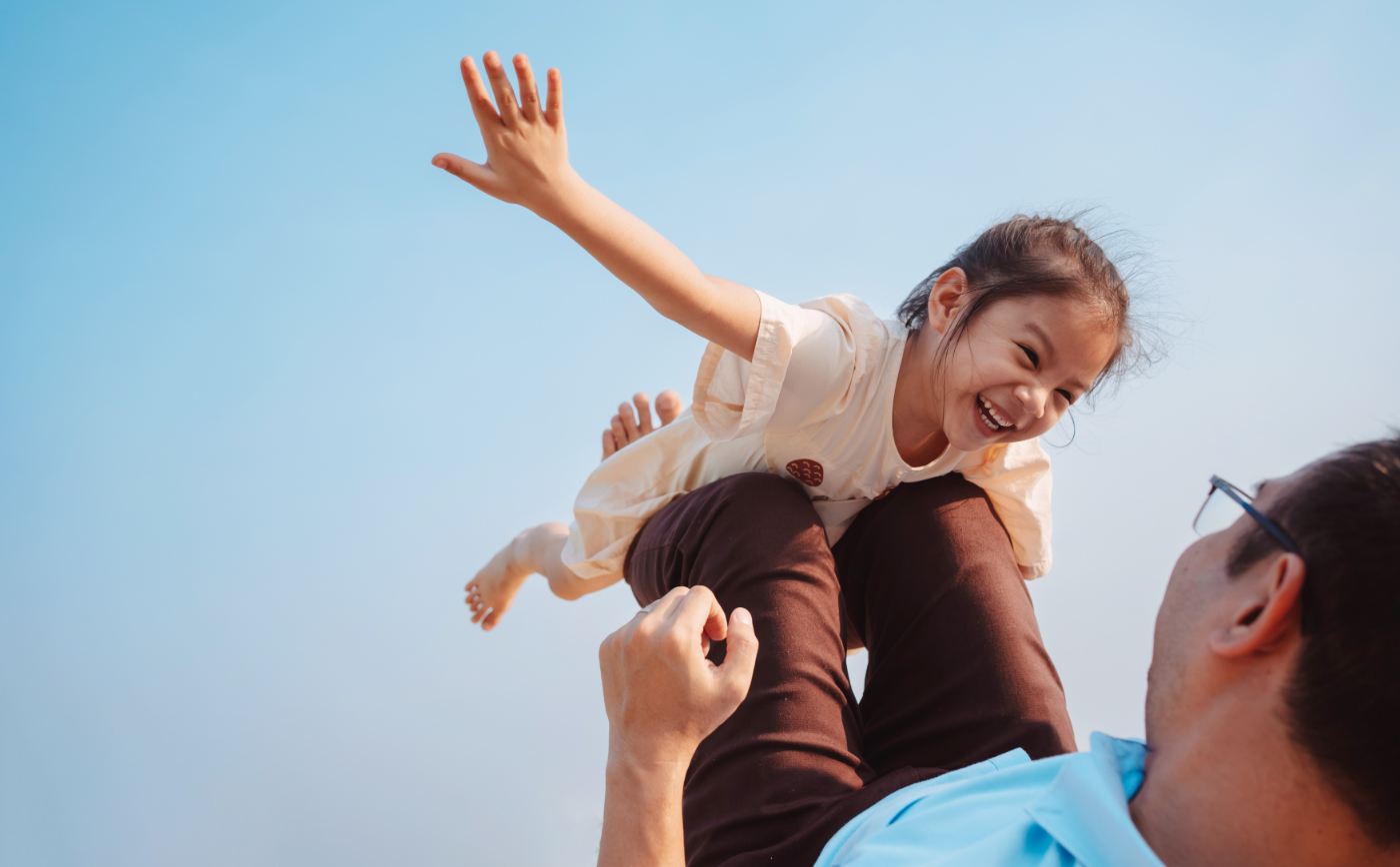 A father lifts his small child in the air like an airplane against a clear blue sky.