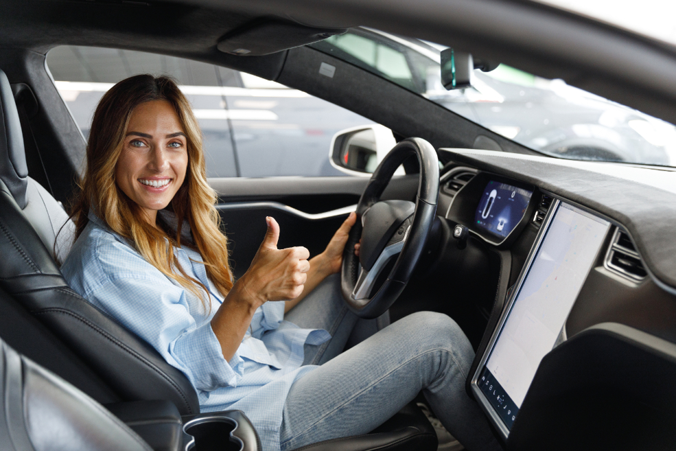 A woman sitting behind the wheel of a car with her seatbelt on, giving a thumbs up