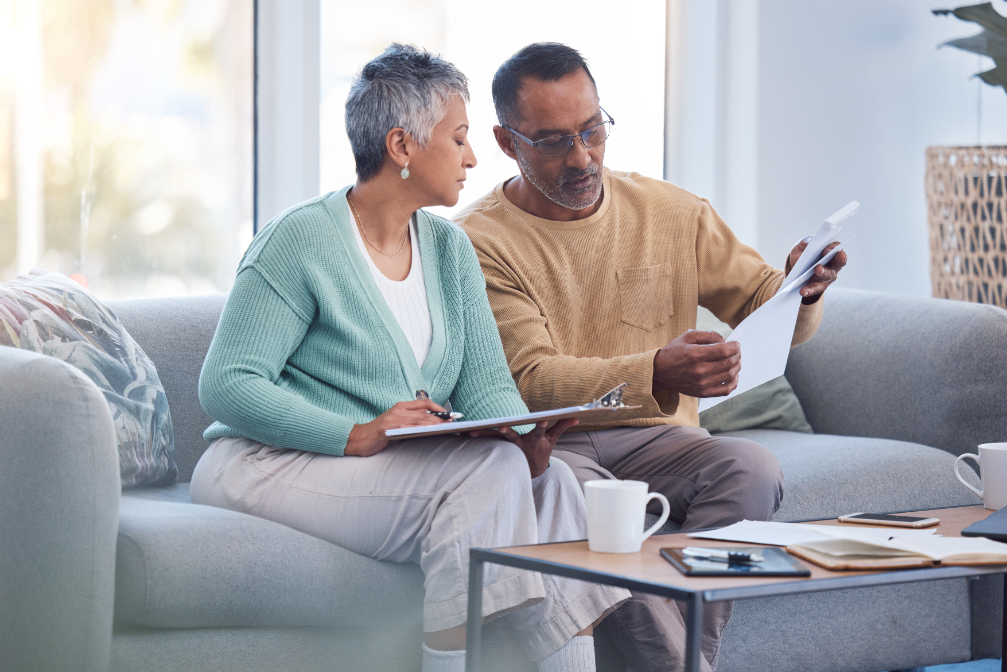 Woman with baby blue cardigan and man with beish crew neck are sitting on a grey couch in a living room space while reading paperwork
