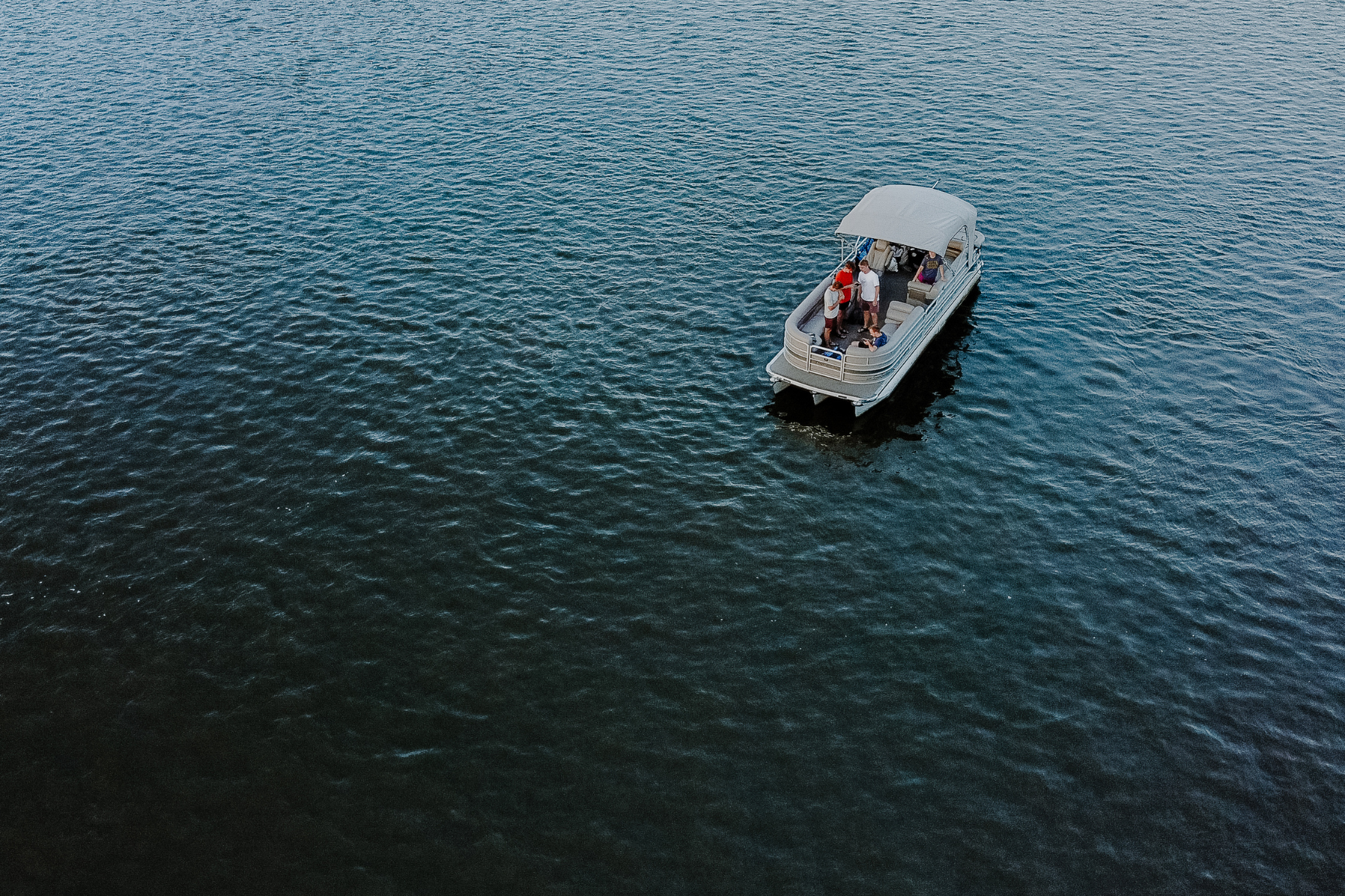 ¿Pontoon boat with people onboard floating on calm, deep blue water.