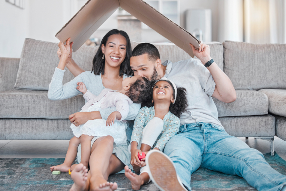 Woman, men and two young girls sitting on rug in front of couch holding a cardboard box over their head