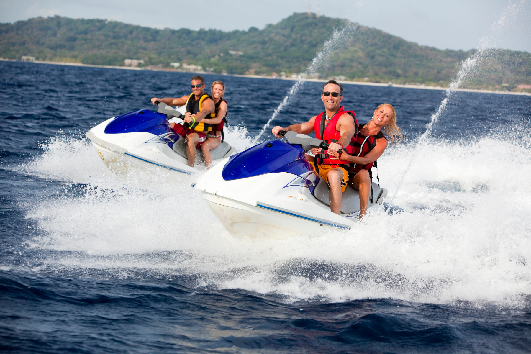 Two couples riding jet skis on the ocean, creating splashes and waves, with a distant shoreline and hills in the background.