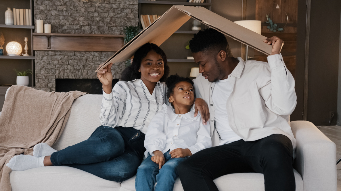 Man, woman and young girl sitting on couch while they hold a cardboard box on their heads.