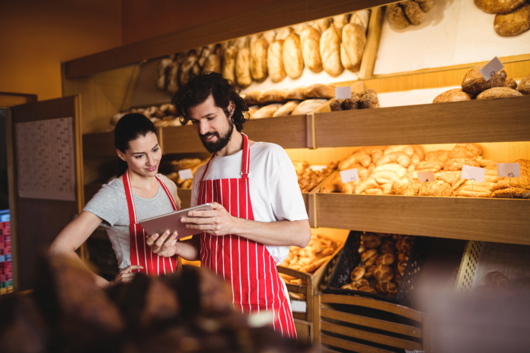 Man and women standing on bakery while checking tablet and wearing red stripped aprons.