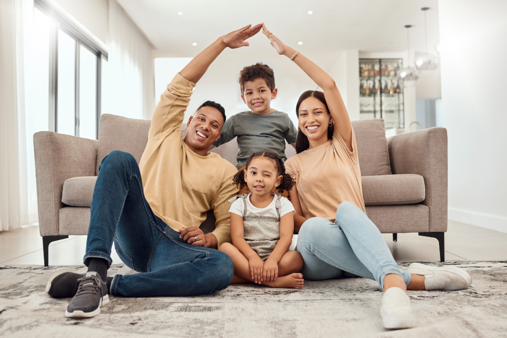 Man,woman,boy and girl sitting on rug in living room. Man and woman touching hands as they form a room figure over the children