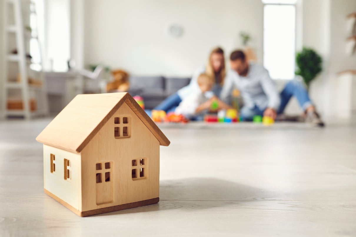 Wooden toy house with blurred family playing on the floor of the living room in the background.
