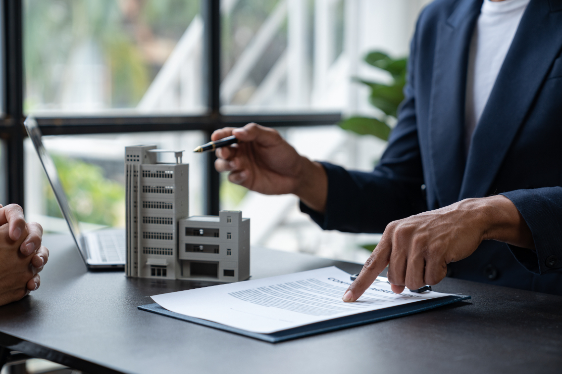 Man wearing suit and pointing at paper on desk with small building toy next to it.