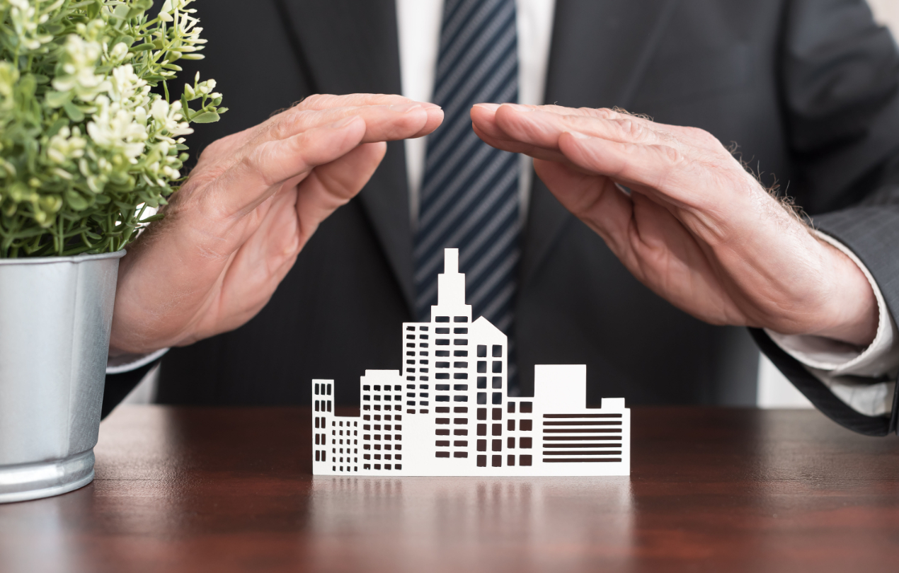 Man wearing suit while sitting down forming a roof shape with hands over small paper buildings.