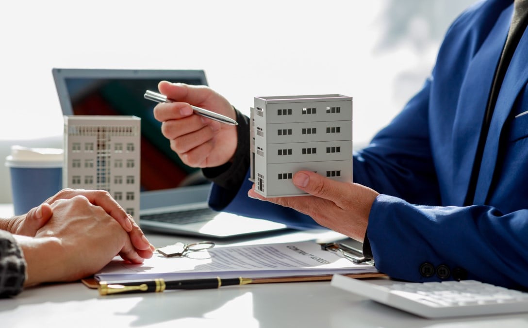Person holding toy house o hand and poiting with pen at it showing another person sitting scross table.