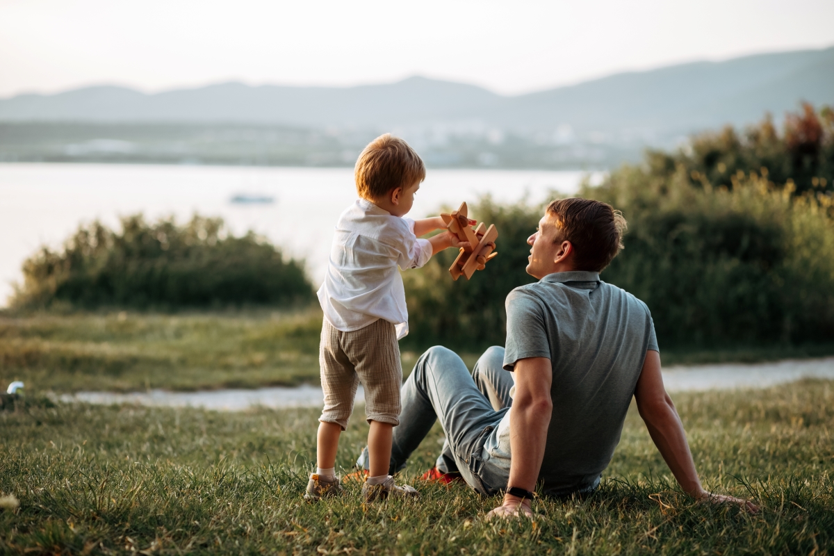 Back view of dad and son sitting on grass with lake view.