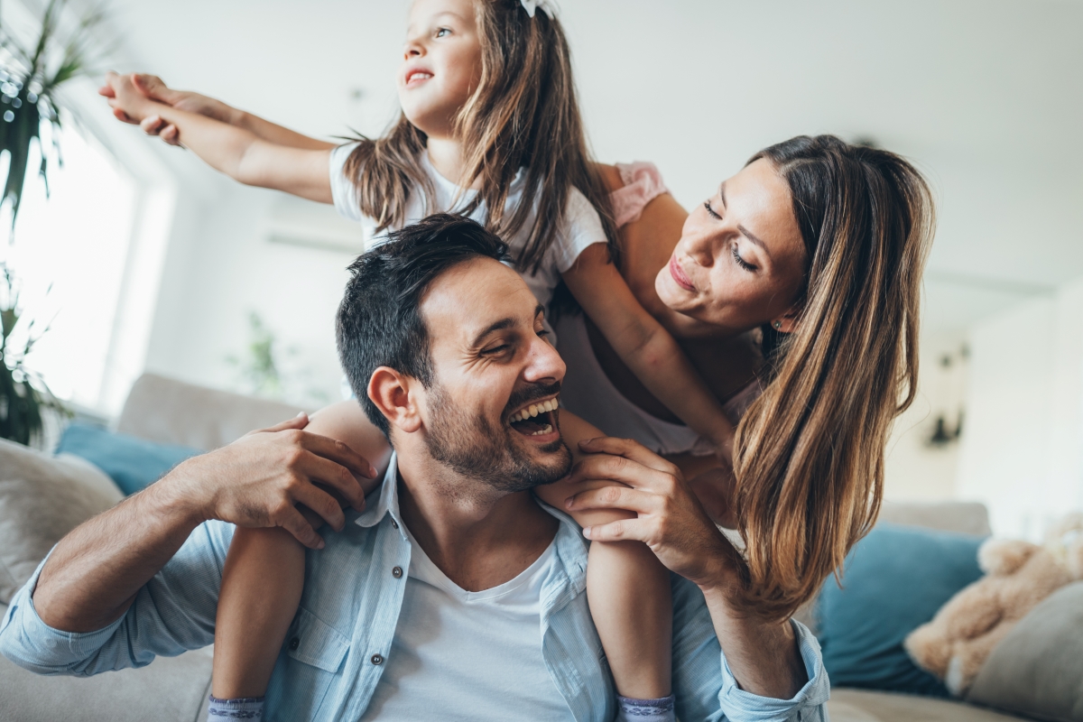 Girl sitting on dads shoulders and mom playing on living room.
