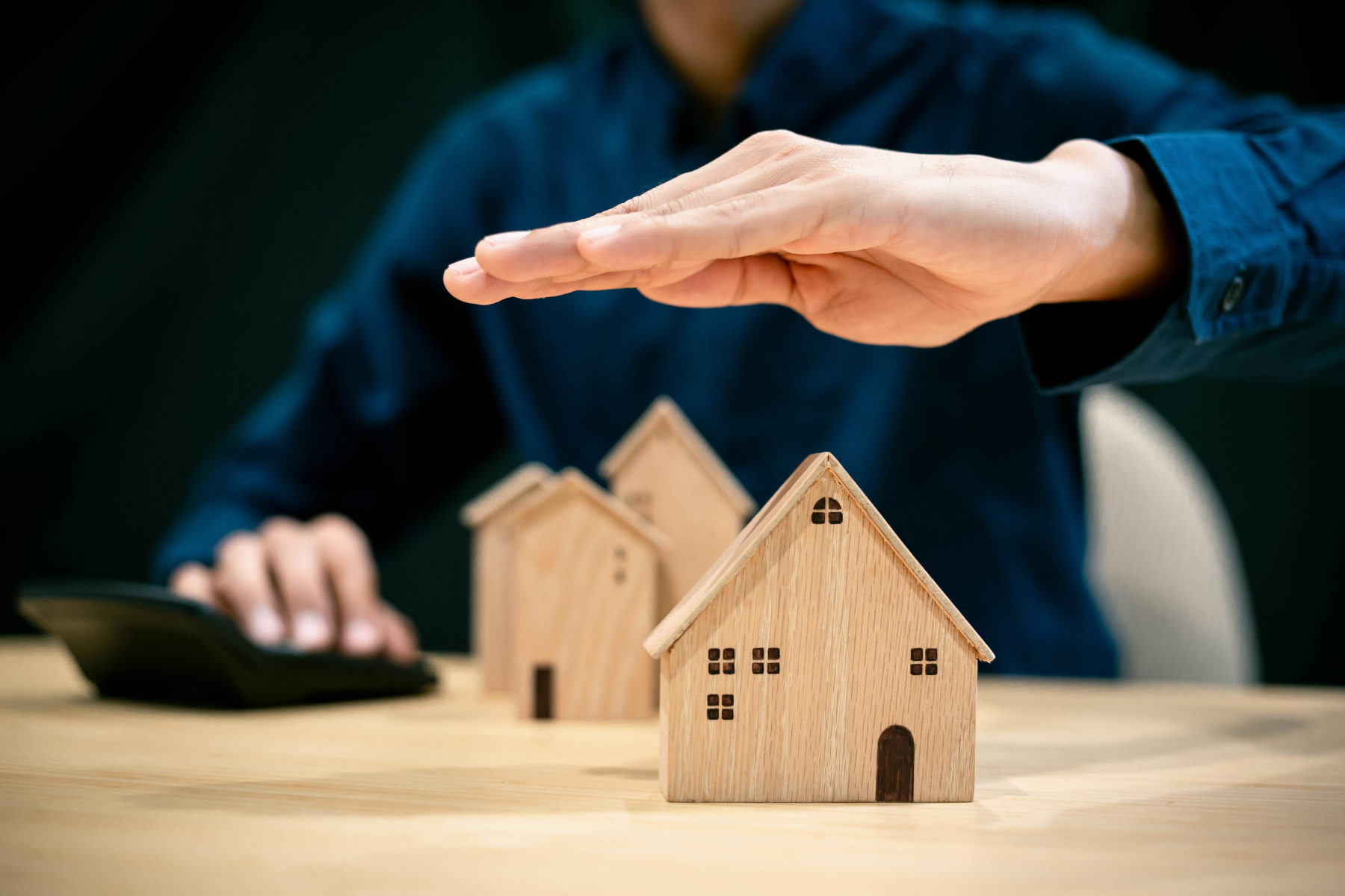 A person in a blue shirt placing their hand protectively over a row of small wooden house models on a table, symbolizing real estate protection or insurance. A calculator is visible on the left side of the table.