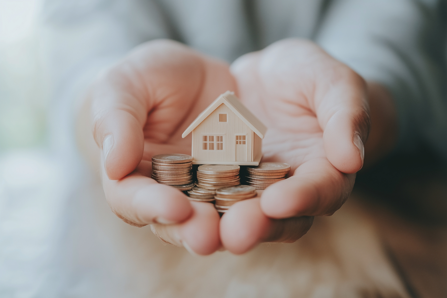 A pair of hands gently holding a stack of coins with a small wooden house model placed on top, symbolizing house insurance.