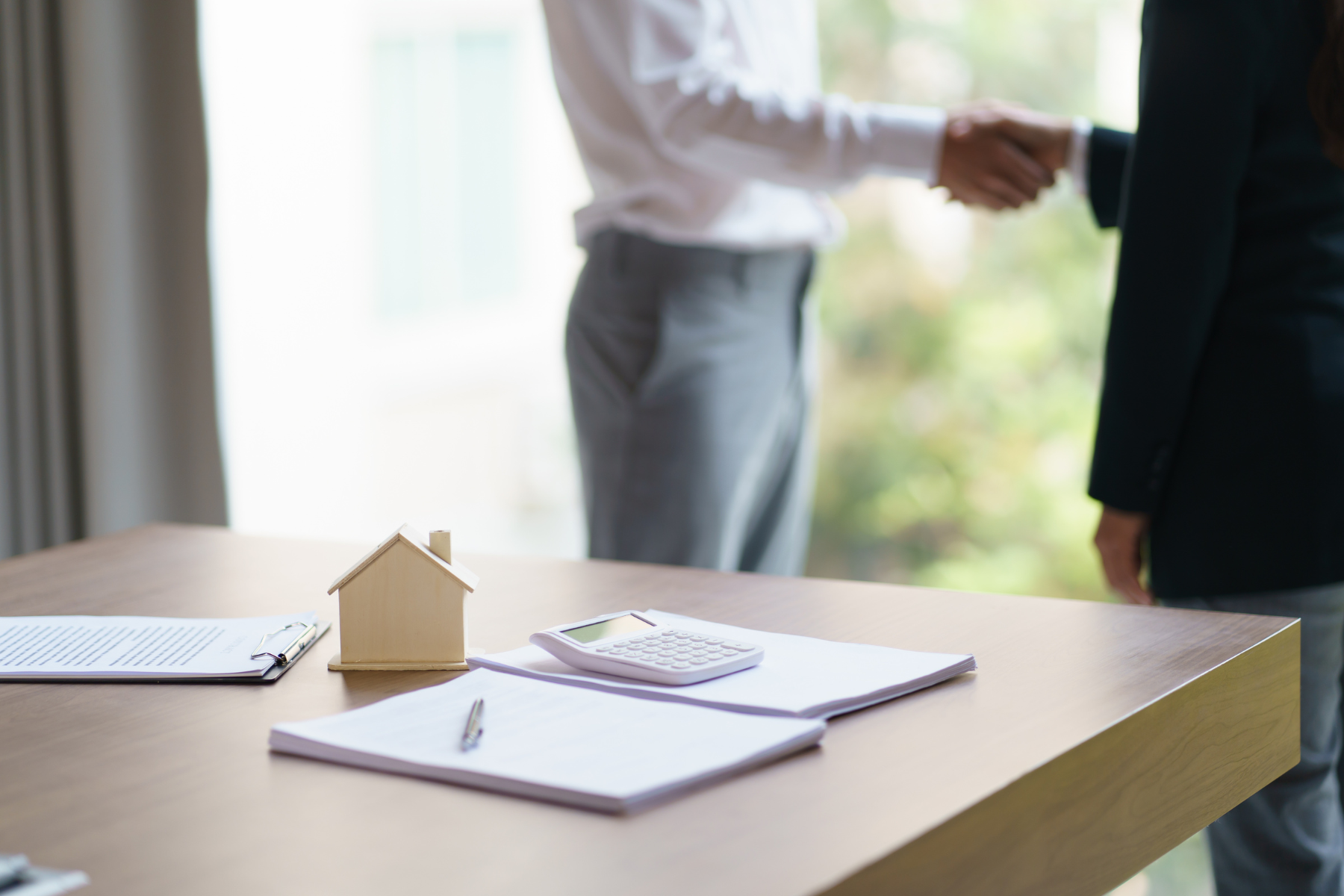 A wooden table with a small house model, documents, a pen, and a calculator in focus. In the blurred background, two people are shaking hands