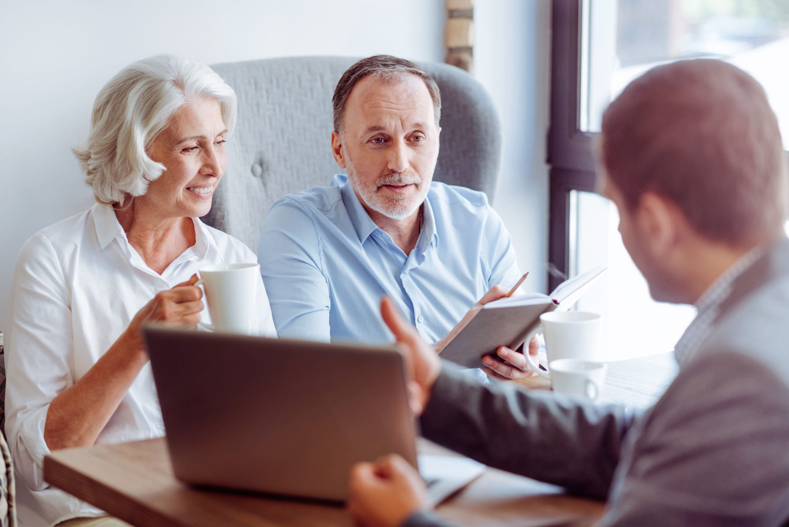 An elderly couple sits at a table with a financial advisor, discussing plans while holding a notebook and a cup of coffee.