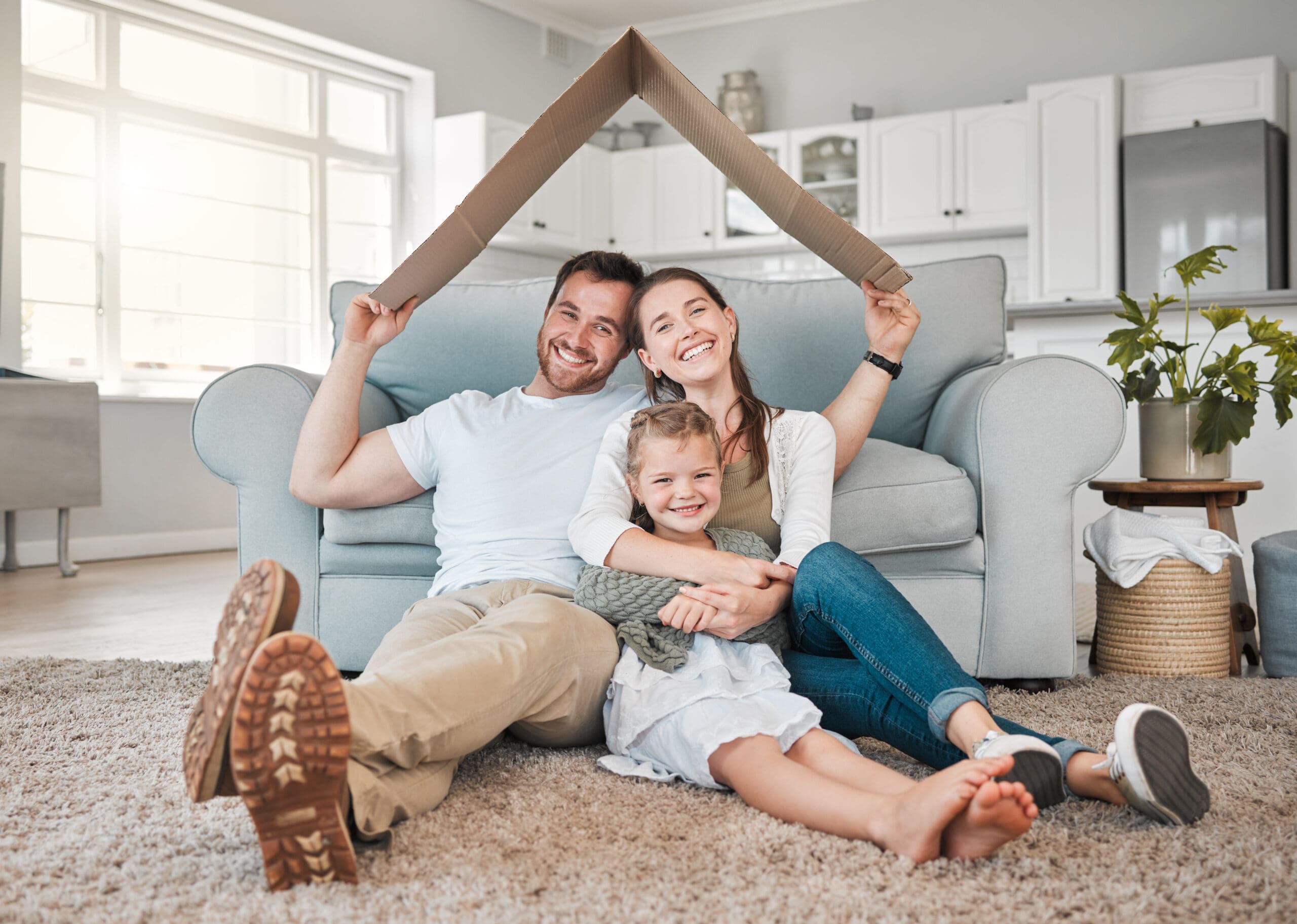 A happy family sits on the floor of their home, smiling and holding a cardboard cutout shaped like a roof above their heads