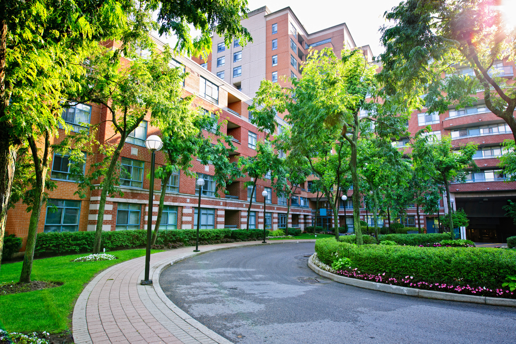 The scene features a red brick residential building surrounded by lush green trees and landscaping. The foreground includes a curved pathway bordered by grass, flowers, and lampposts, creating a serene and inviting atmosphere. The bright sunlight filters through the trees, adding warmth to the scene.