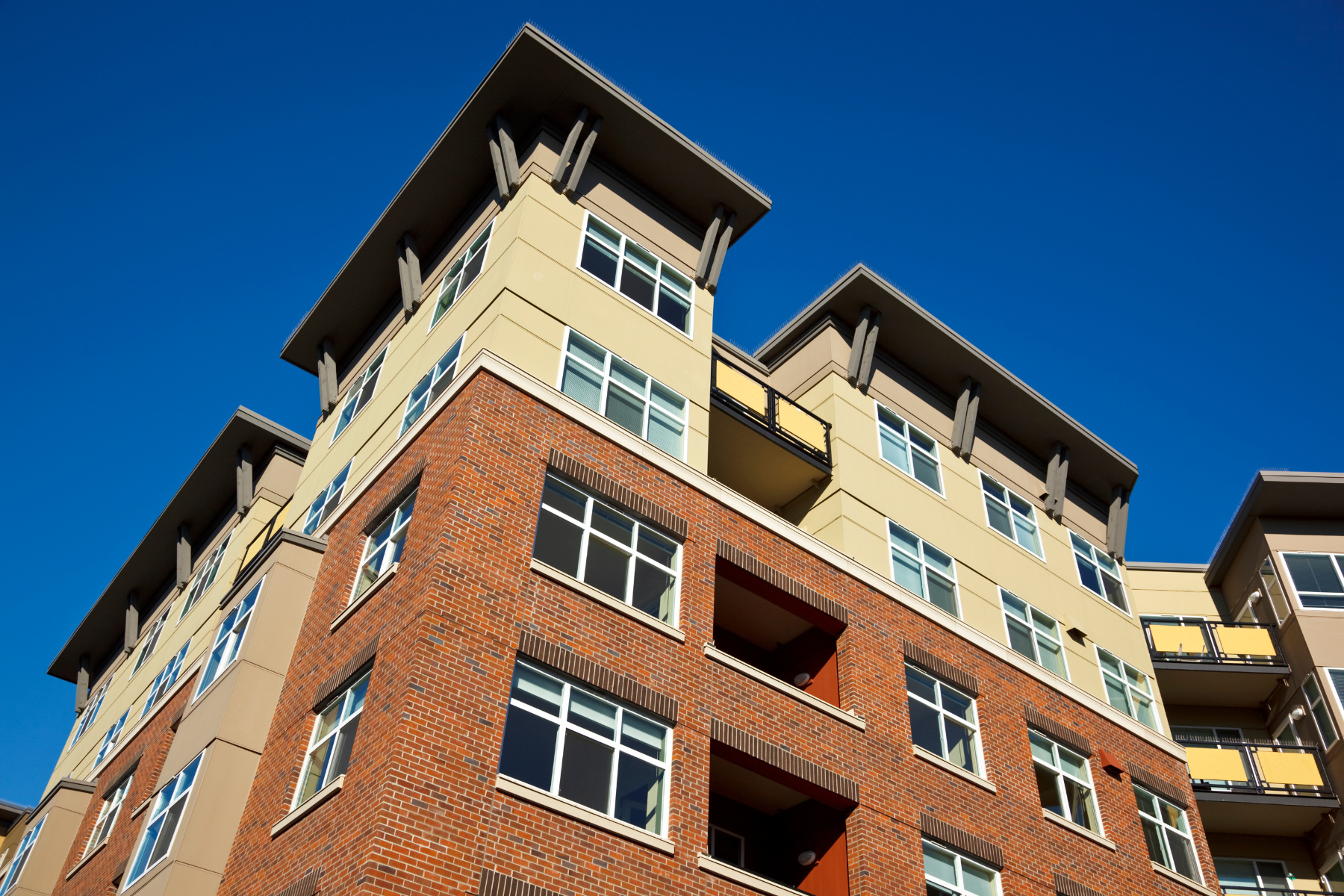 The photograph shows a multi-story residential building with modern architecture. The lower portion of the building features a red brick façade, while the upper levels are finished in beige and tan tones. The structure includes many windows, with some balconies visible. The background is a clear blue sky, emphasizing the bright and sunny weather.