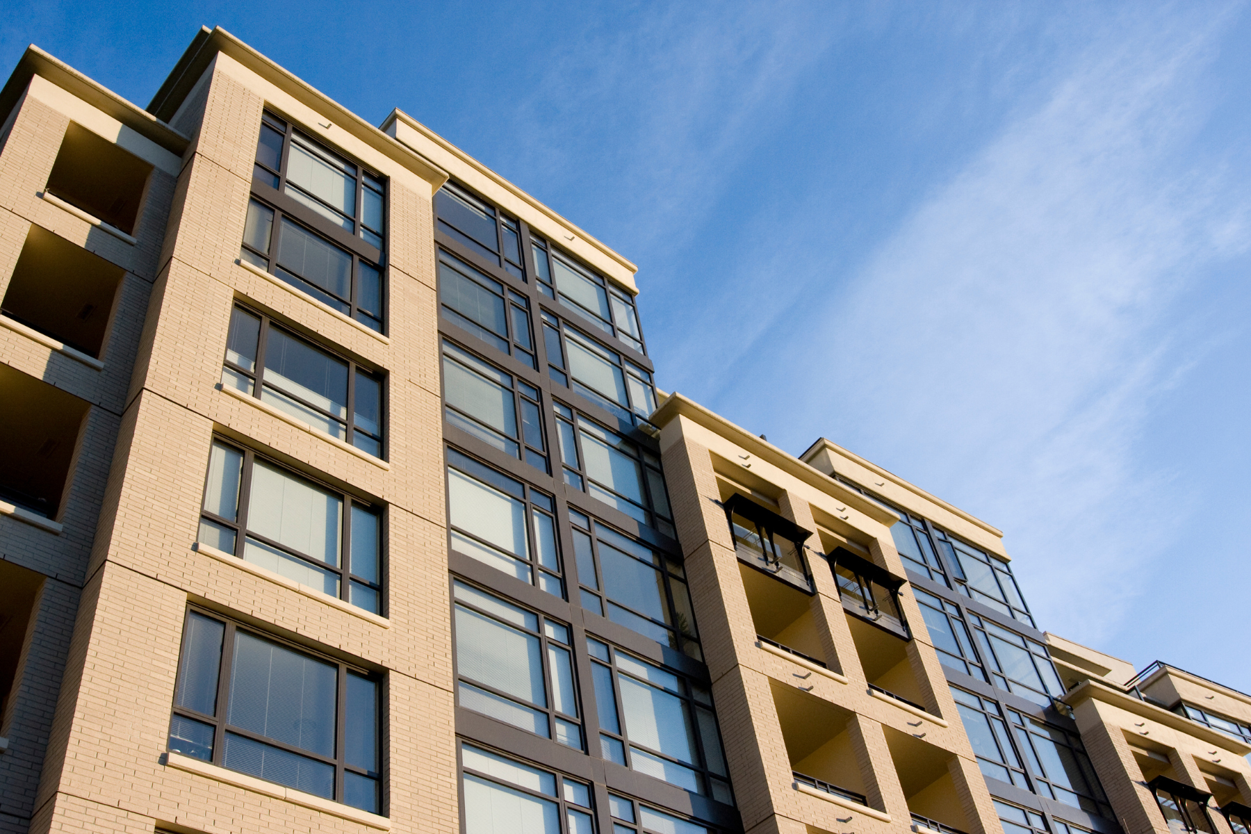This is an image of a contemporary building with beige brick walls and large glass windows. The building's design includes balconies with metal railings and a geometric structure. The sky in the background is a clear blue with light clouds.