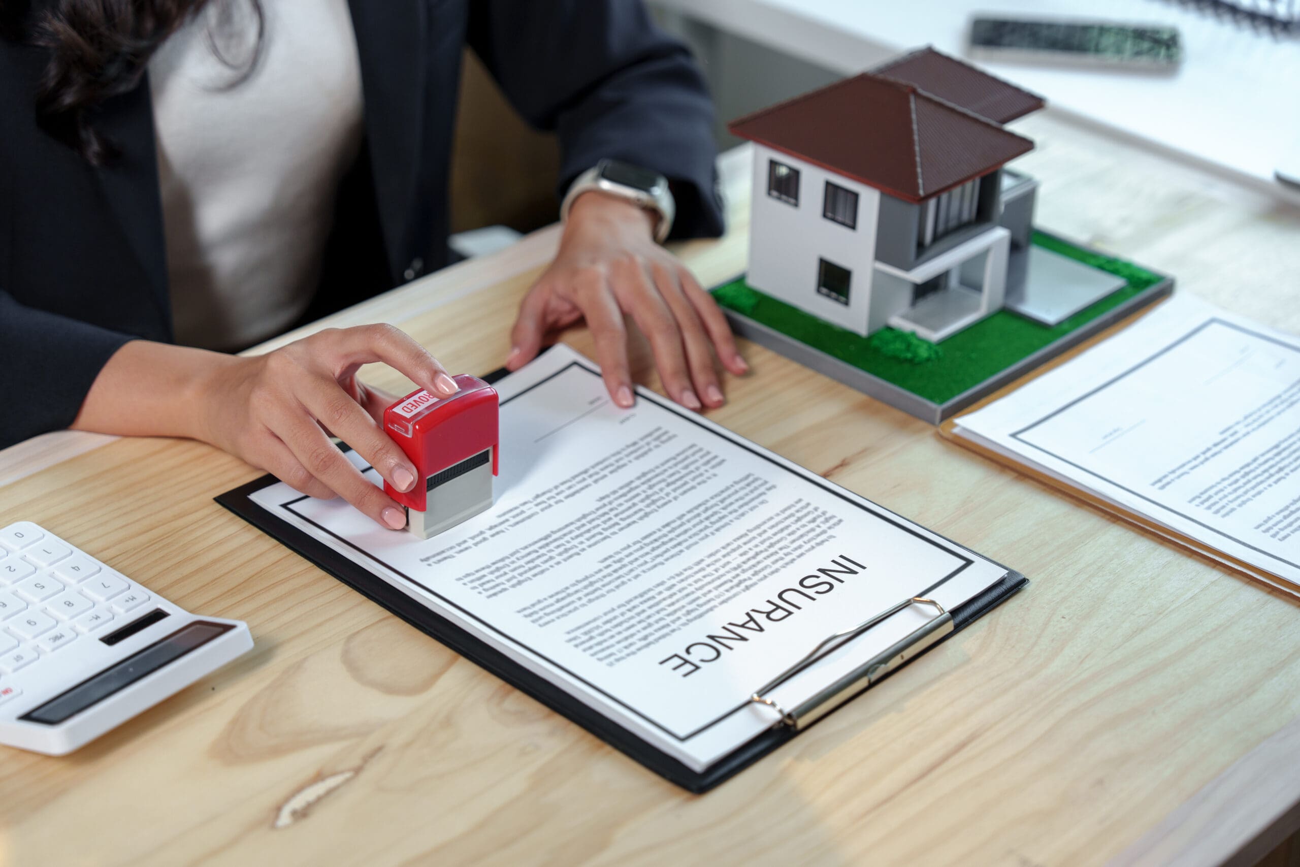 A person presses a red stamp marked “APPROVED” onto a document labeled "INSURANCE" on a clipboard. A model house, additional papers, and a calculator are also on the desk.