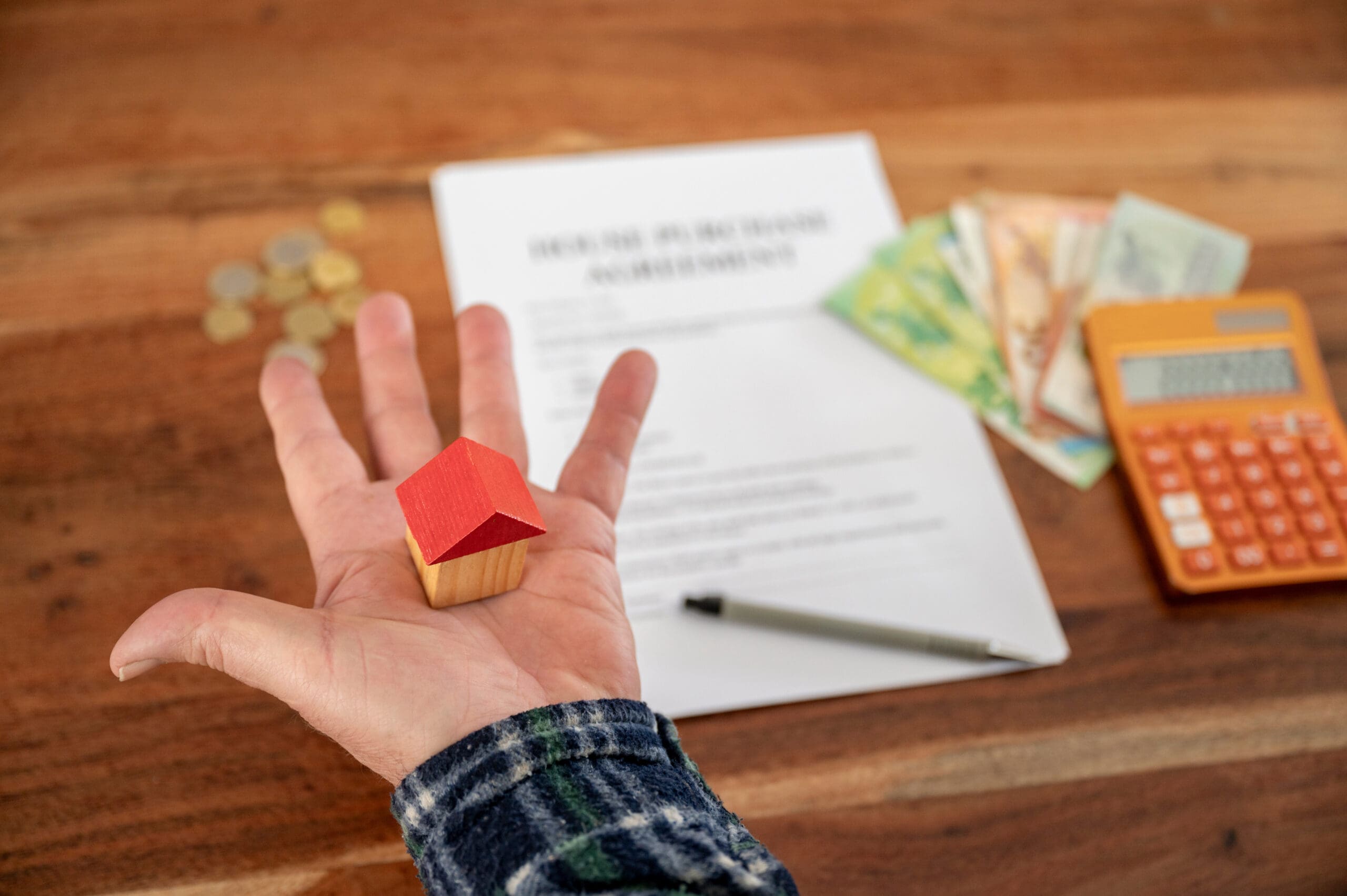 A hand holds a small wooden model of a house. In the background on a wooden surface are a house purchase agreement document, paper currency, coins, a calculator, and a pen, slightly out of focus.