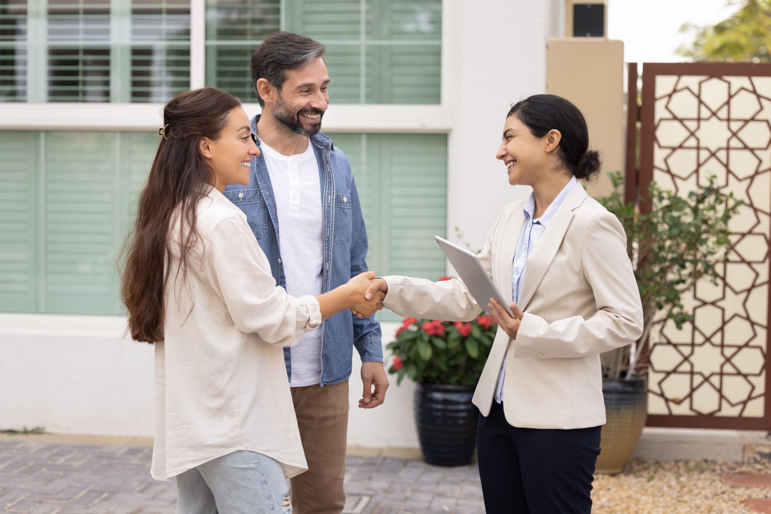 A woman and a man stand outside a house, smiling as the woman shakes hands with another woman holding a tablet, likely in a professional or real estate role.