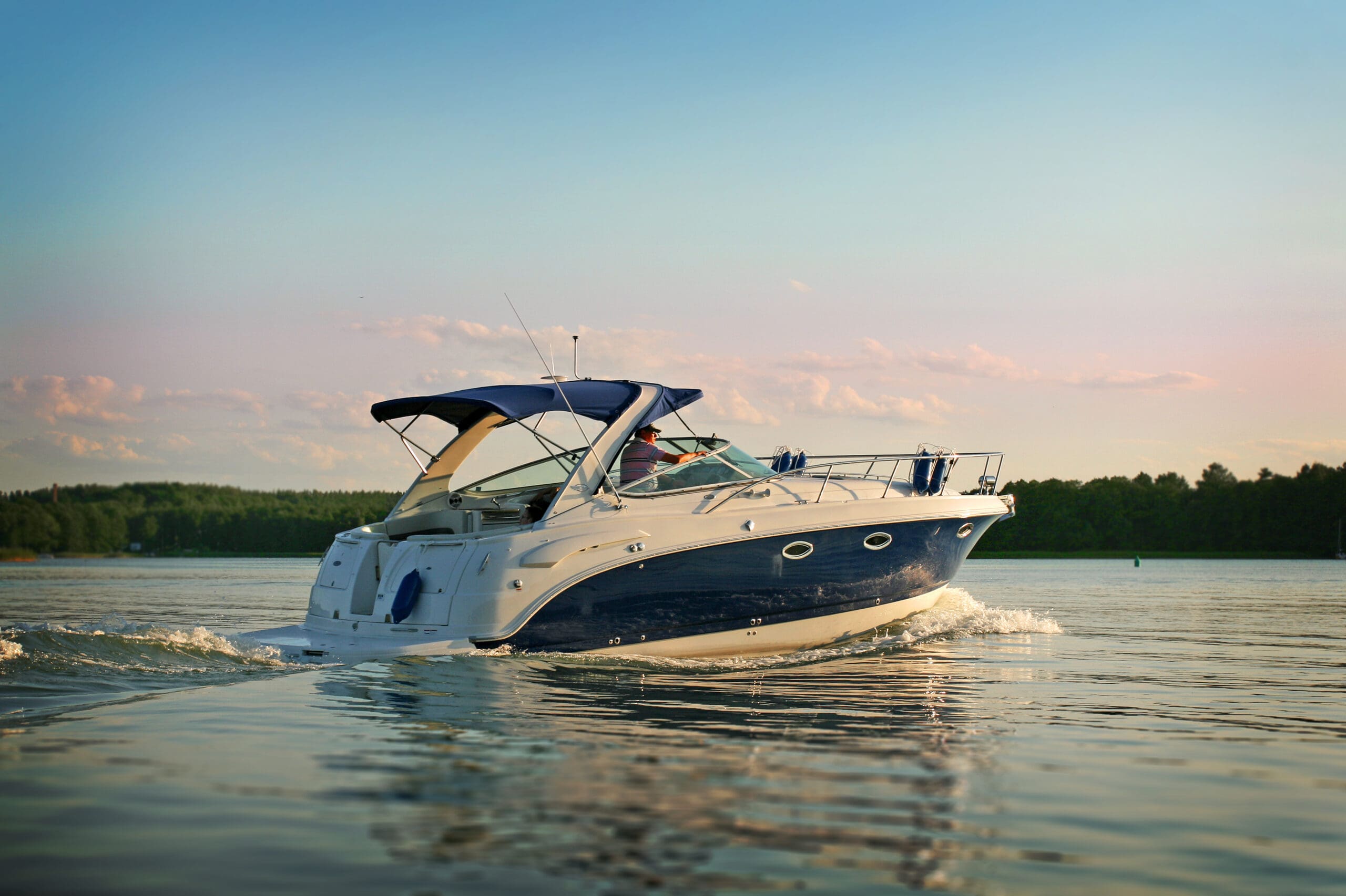The image shows a boat moving across a calm body of water. The boat is white with a dark blue lower section and a blue canopy covering the front area. A person is visible under the canopy, wearing a hat and a light-colored shirt, steering the boat. The water around the boat has gentle waves, reflecting the boat's image. In the background, there is a green forested shoreline on the left and right, with a light blue sky above that transitions to pink near the horizon, indicating sunset. A small green buoy floats in the distance on the right side of the image.