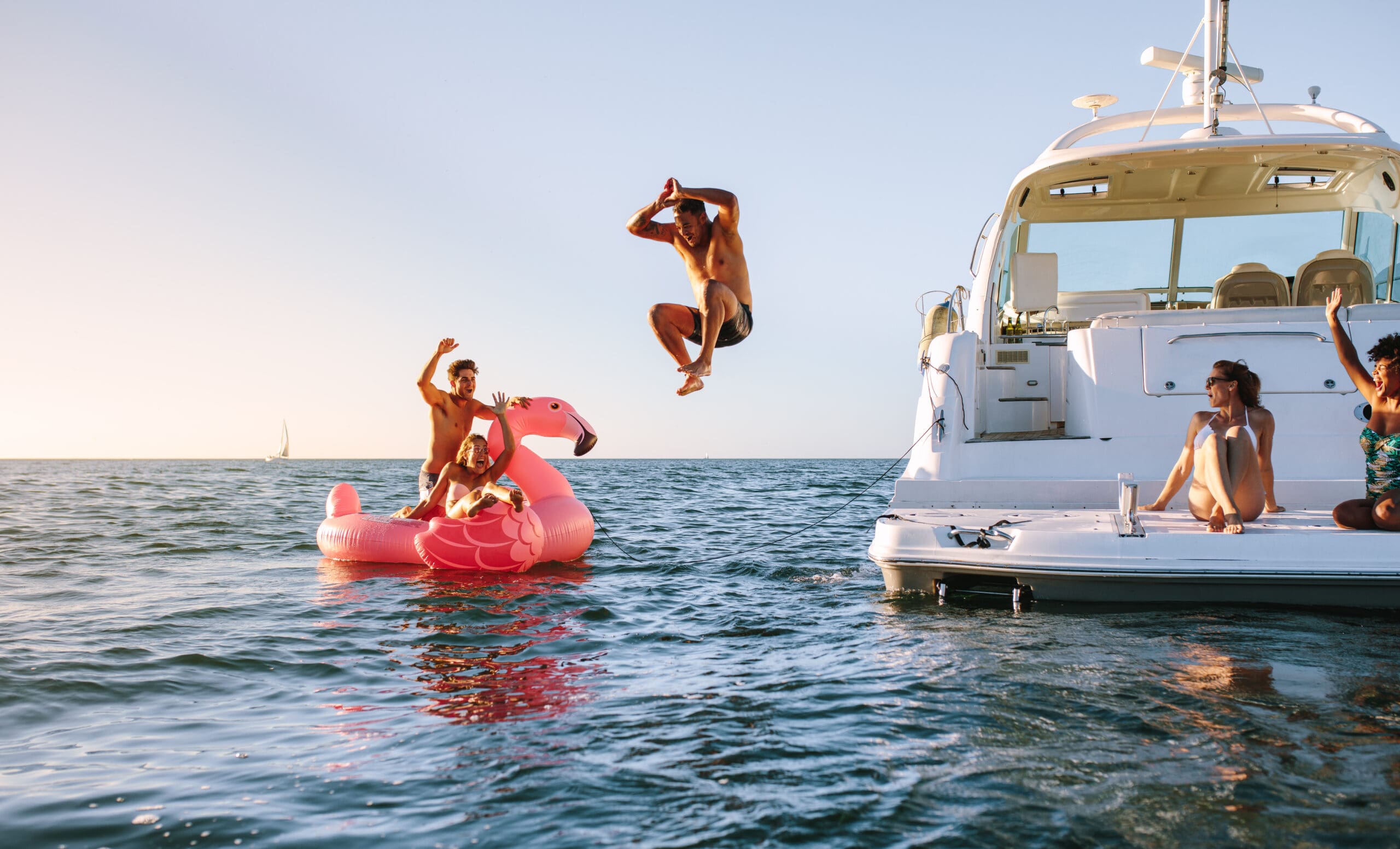 This image features a group of people having fun at sea. One person is mid-jump, diving into the water from a boat. Another person is riding an inflatable pink flamingo, and a few others are seated on the boat, enjoying the moment.