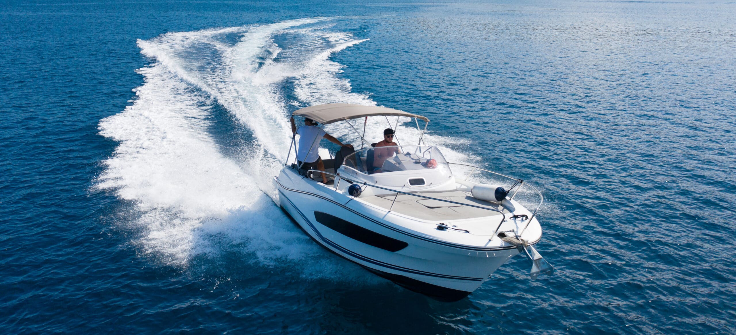 This image shows a boat speeding through the clear blue water, creating a wake behind it. There are two people aboard the boat, with one steering the boat, while the other seems to be enjoying the ride in the sun.
