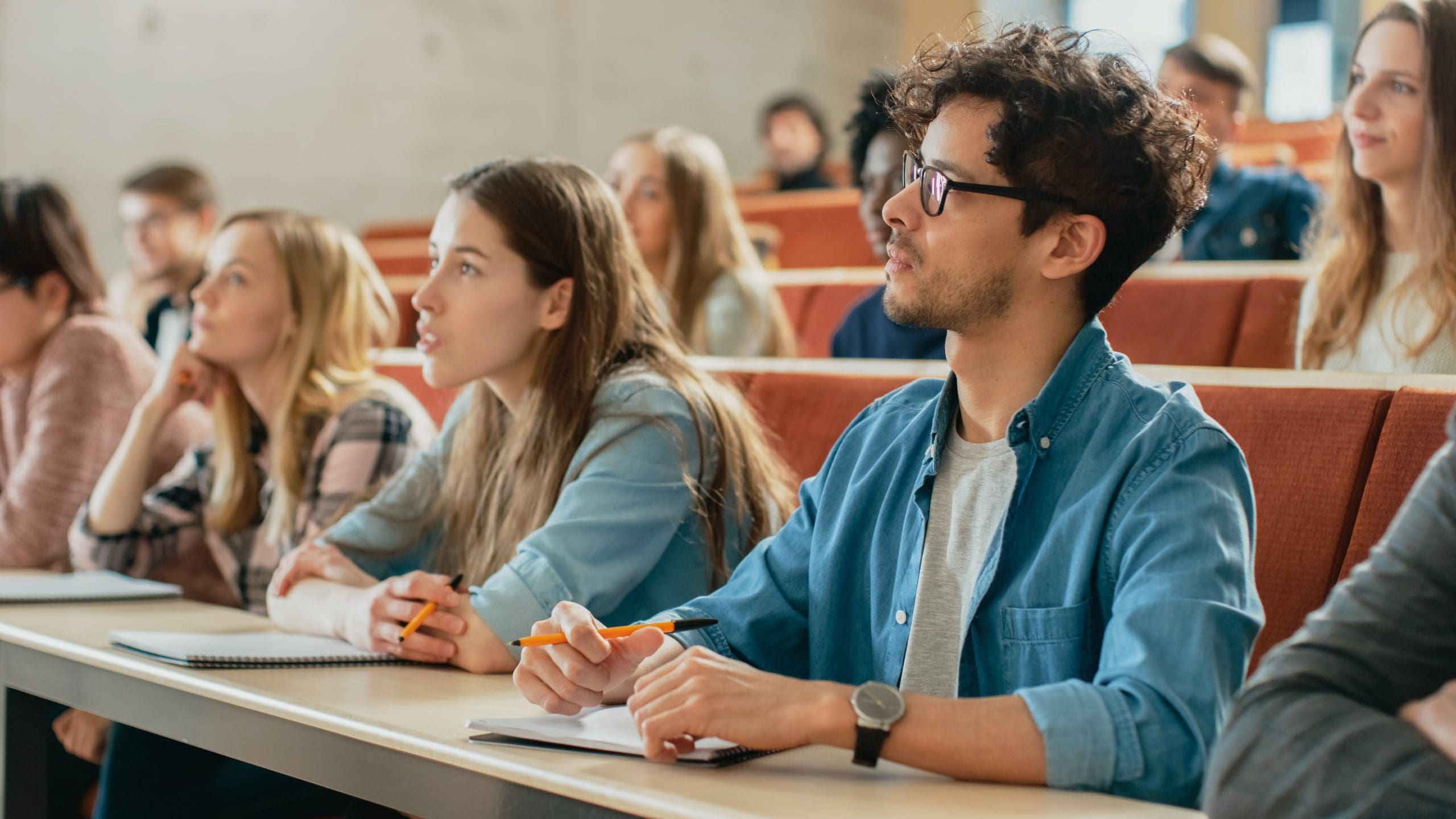A group of students seated in a lecture hall, attentively listening and taking notes.