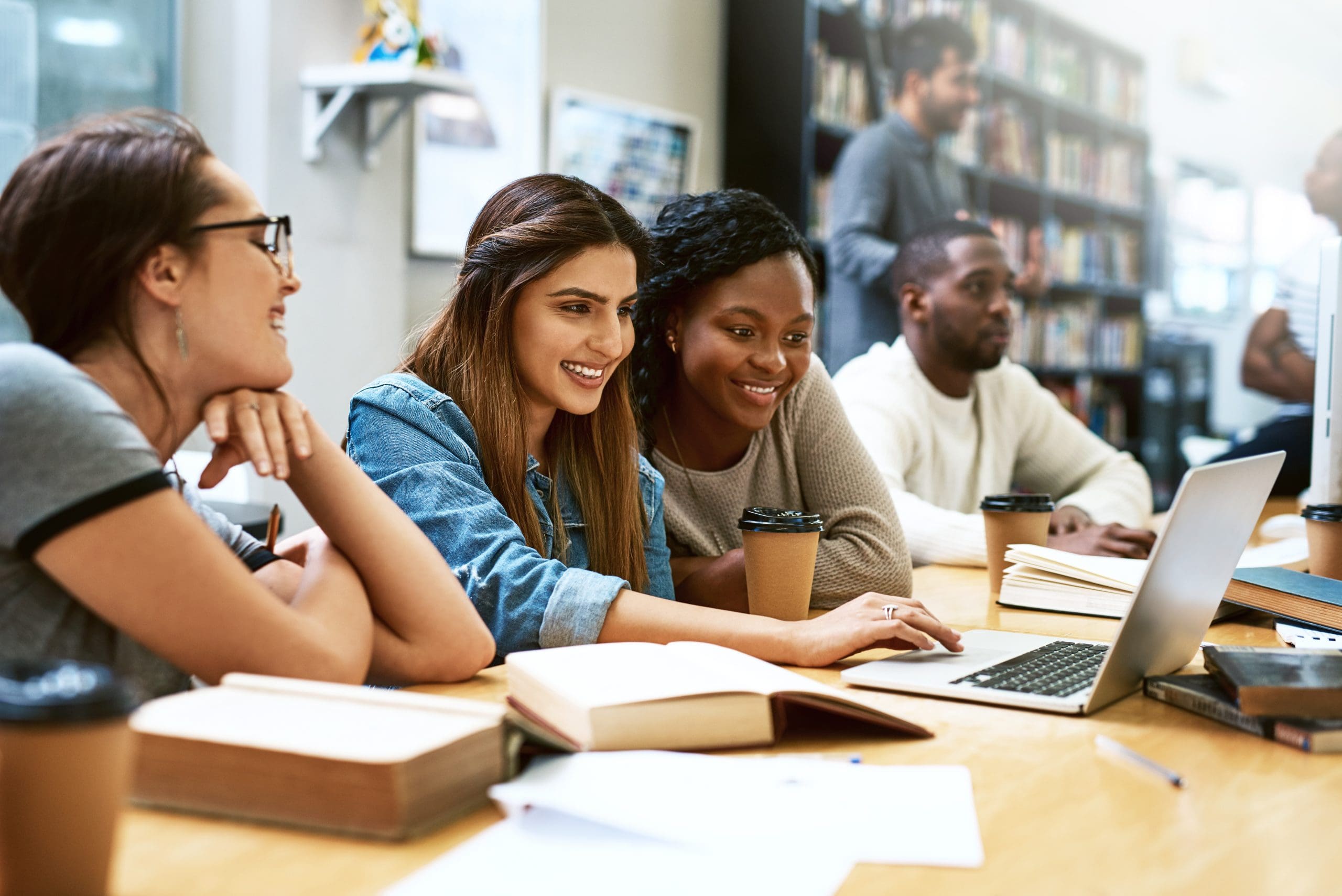 Several students gathered around a laptop in a library or study space, smiling and looking at the screen with books and coffee cups nearby.
