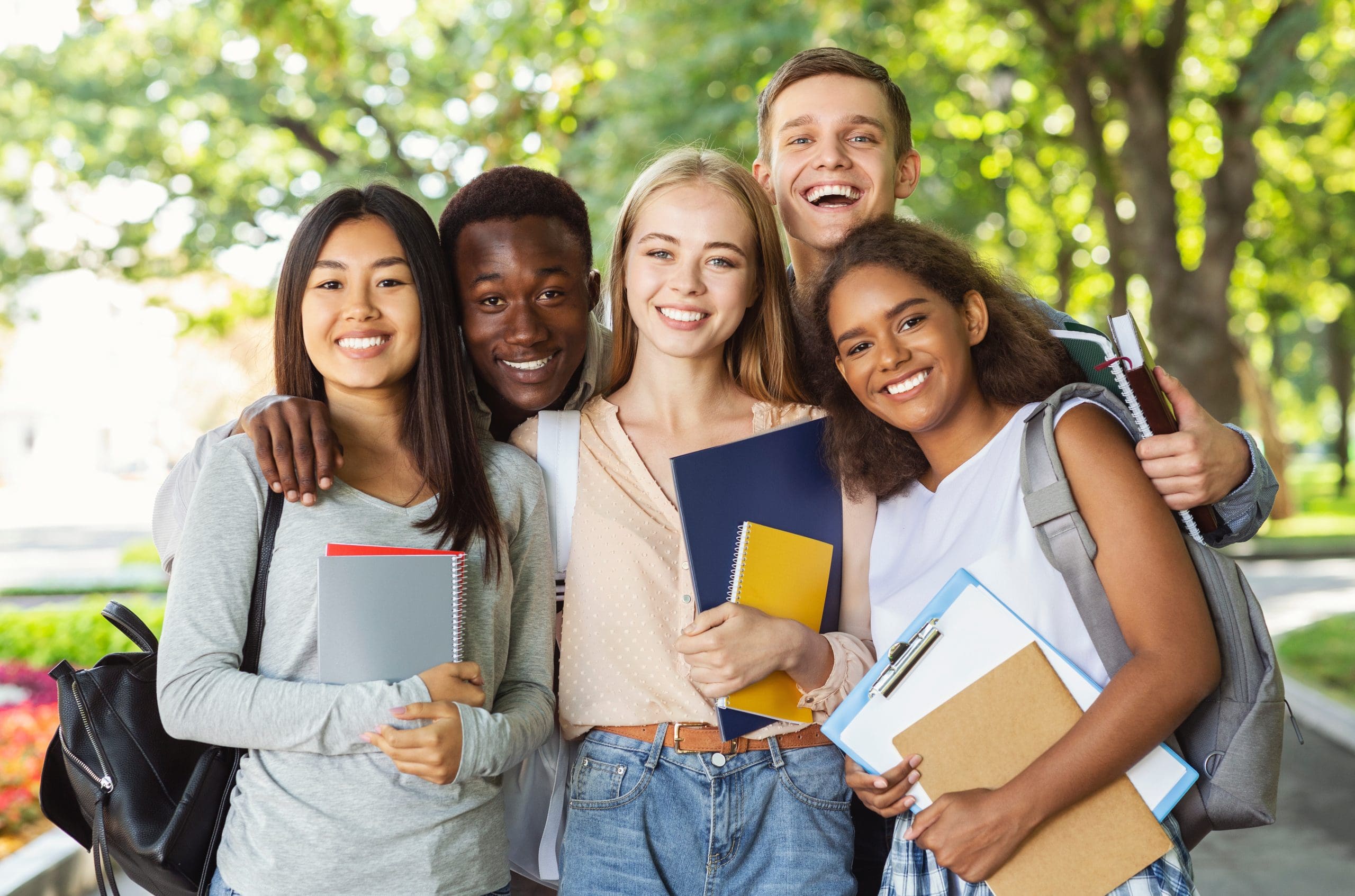 Five smiling students standing together outside in a park-like area, holding notebooks and backpacks.
