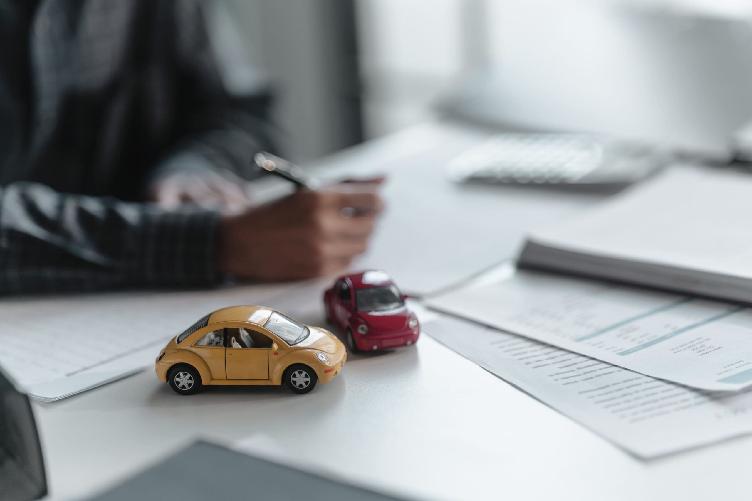 Two toy cars on desk. Person signing papers next to them. Car insurance concept.