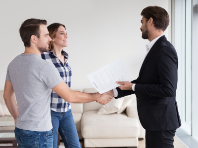 A man and a woman stand in a living room shaking hands with a man in a dark suit who is holding a document. A white sofa and bright windows are visible in the background.
