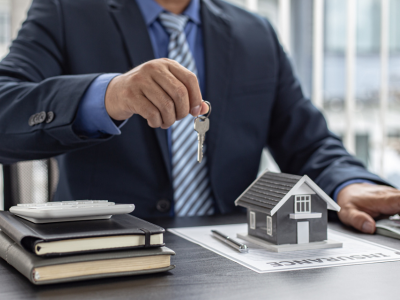 A man in a navy suit and striped tie sits at a desk holding a set of keys. A small gray house model, a document labeled "Insurance," a calculator, and stacked notebooks are arranged on the desk in front of him.