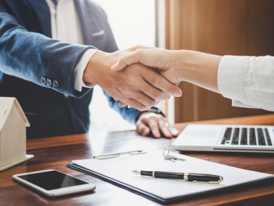 A close-up of two people shaking hands across a wooden desk. A clipboard with paperwork, a pen, keys, a smartphone, a laptop, and a small wooden house model are visible on the desk surface.